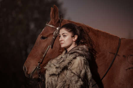 A young brunette woman in a gray coat poses with a brown horse. Cold autumn in the forest on the...