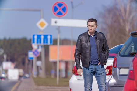 A young adult man in a black leather jacket and jeans walks along a city street near cars on a sunny...