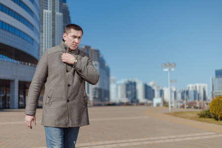 A young adult man in a gray coat walks along a city street on a sunny day in springの写真素材