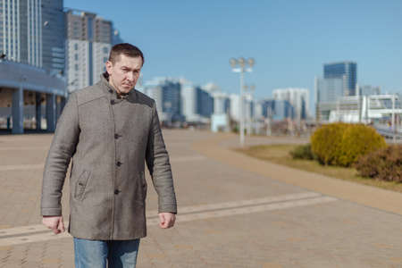 A young adult man in a gray coat walks along a city street on a sunny day in spring