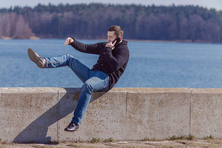 Young adult man in black knitted jacket and jeans sitting talking on a smartphone on the edge on the waterfront near the lake on a sunny dayの写真素材