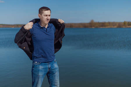 A young adult man in a black knitted jacket and jeans stands on the promenade near the lake on a sunny dayの写真素材