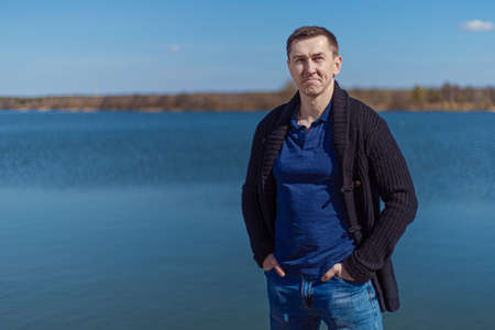 A young adult man in a black knitted jacket and jeans stands on the promenade near the lake on a...