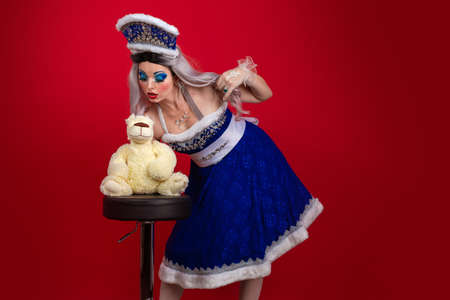 Emotional Snow Maiden in an elegant bright blue suit with a crown posing with a big teddy bear on a red backgroundの写真素材