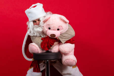 Emotional Santa Claus with a long white beard in a red coat and white hat posing with a big teddy...