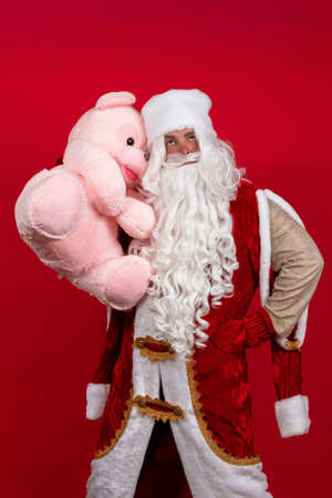 Emotional Santa Claus with a long white beard in a red coat and white hat posing with a big teddy pink bear.の写真素材
