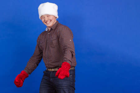 Young guy in a red cap and mittens Santa Claus poses on a blue backgroundの写真素材