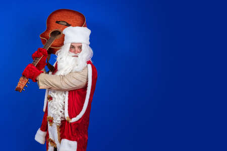 Emotional Santa Claus with a long white beard in a red coat and white hat sings and plays the guitar on a blue backgroundの写真素材