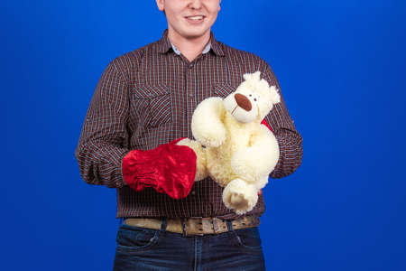 Young man in a red cap and mittens Santa Claus holds a teddy bear in his hands and poses on a blue backgroundの写真素材