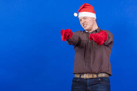 Young guy in a red cap and mittens Santa Claus poses on a blue backgroundの写真素材