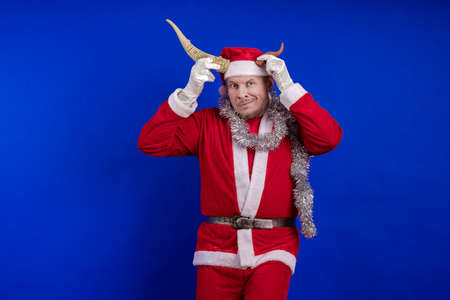 Male actor in a costume of Santa Claus holds horns, jokes, grimaces and poses on a blue background