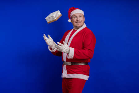 Emotional male actor in a costume of Santa Claus holds a box with a gift in his hands, juggles and...