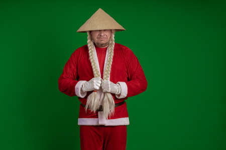 Male actor dressed as Santa Claus with long braids hairstyle and Asian hat posing on green chrome backgroundの写真素材