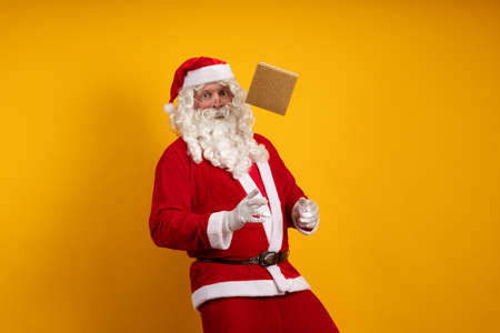Santa Claus holds a box with a gift in his hands, juggles and poses on a yellow background