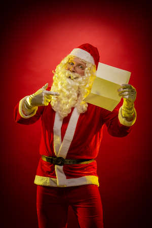 Santa Claus holds a sheet of paper in his hands a Christmas letter reads and poses on a dark red backgroundの写真素材