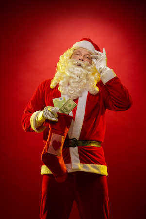 Male actor in a suit of Santa Claus holds in his hands banknotes, money and a Christmas gift sock and poses on a dark red backgroundの写真素材