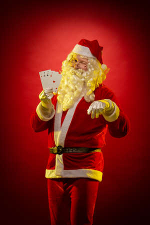 Santa Claus holds playing cards in his hands and poses on a dark red background