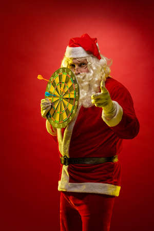 Santa Claus holds a darts target in his hands and poses on a dark red backgroundの写真素材