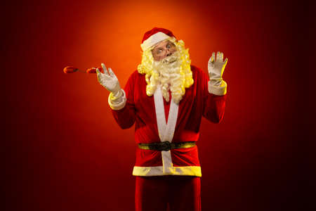 Male actor in a costume of Santa Claus holds red fur handcuffs and a whip in his hands and poses on a dark red backgroundの写真素材