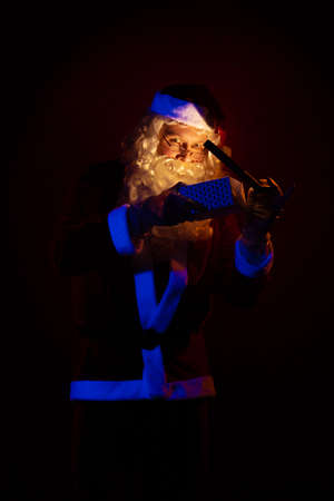 Male actor dressed as Santa Claus holds a gift box in his hands, looks into it and poses on a yellow backgroundの写真素材