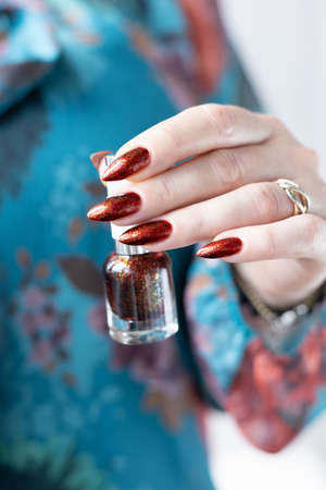 Female hand with long nails and golden brown maroon, manicure holds a bottle of nail polish