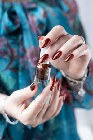 Female hand with long nails and golden brown maroon, manicure holds a bottle of nail polish