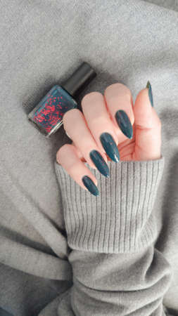 Female hand with long nails and dark blue teal manicure holds a bottle of nail polish