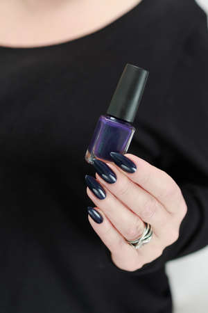 Female hand with long nails and dark purple blue manicure holds a bottle of nail polish