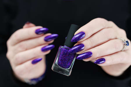 Female hand with long nails and purple plum manicure holds a bottle of nail polish