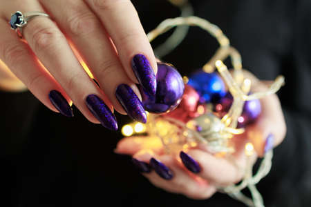 Female hands with long nails and bright purple and blue manicure hold a gift box and Christmas balls. Congratulations to the New Year and Christmas.の写真素材