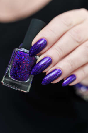 Female hand with long nails and purple plum manicure holds a bottle of nail polish