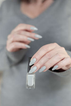 Female hand with long nails and a gray manicure holds a bottle of nail polishの写真素材
