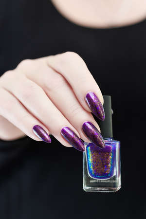 Female hand with long nails and purple plum manicure holds a bottle of nail polish