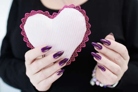 Female hands with purple long nails hold a pink toy soft heartの写真素材