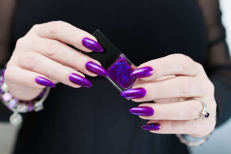 Female hand with long nails and purple plum manicure holds a bottle of nail polishの写真素材