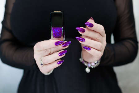 Female hand with long nails and purple plum manicure holds a bottle of nail polishの写真素材