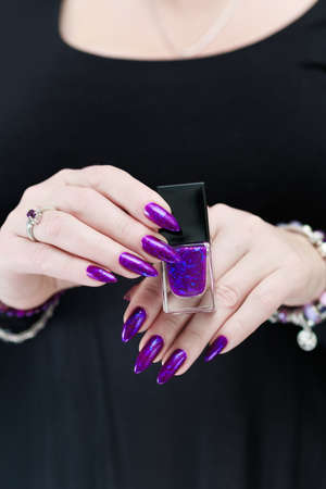 Female hand with long nails and purple blue manicure holds a bottle of nail polish