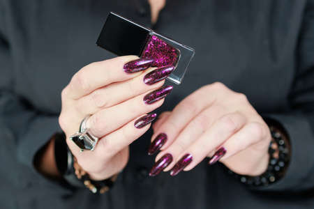 Female hand with long nails and dark purple burgundy manicure holds a bottle of nail polish