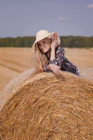 A blonde girl with long hair in a white hat is resting and posing near the sheaves of hay in a field on a sunny dayの写真素材
