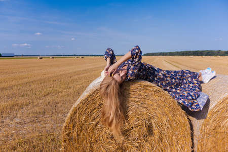 A blonde girl with long hair in a white hat is resting and posing near the sheaves of hay in a field on a sunny dayの写真素材
