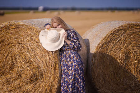 A blonde girl with long hair in a white hat is resting and posing near the sheaves of hay in a field...