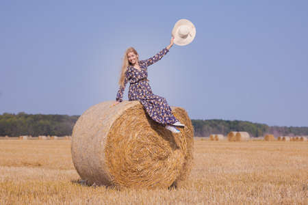 A blonde girl with long hair in a white hat is resting and posing near the sheaves of hay in a field on a sunny dayの写真素材