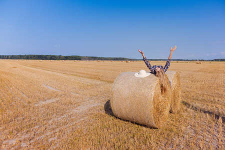 A blonde girl with long hair in a white hat is resting and posing near the sheaves of hay in a field on a sunny dayの写真素材