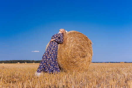 A blonde girl with long hair in a white hat is resting and posing near the sheaves of hay in a field on a sunny dayの写真素材