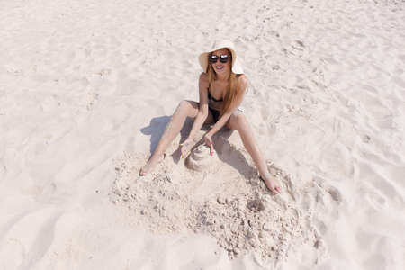 A blonde girl with long hair in a white hat is resting on the white sand near the sea on a sunny dayの写真素材