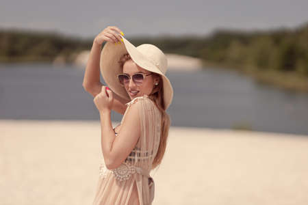 A blonde girl with long hair in a white hat is resting on the white sand near the sea on a sunny dayの写真素材