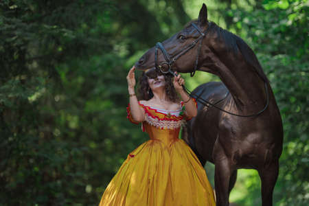 A young woman in a vintage yellow dress walks with a brown horse in a green park on a summer dayの写真素材