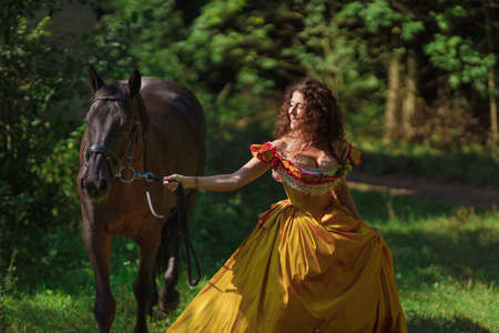 A young woman in a vintage yellow dress walks with a brown horse in a green park on a summer day