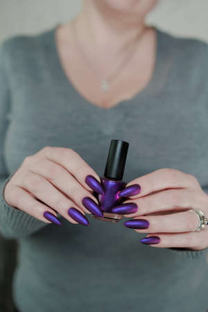Female hand with long nails and purple plum manicure holds a bottle of nail polishの写真素材