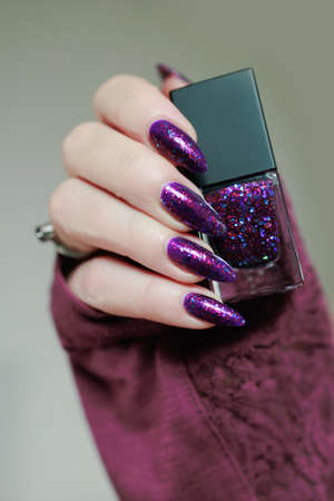 Female hand with long nails and purple plum manicure holds a bottle of nail polish